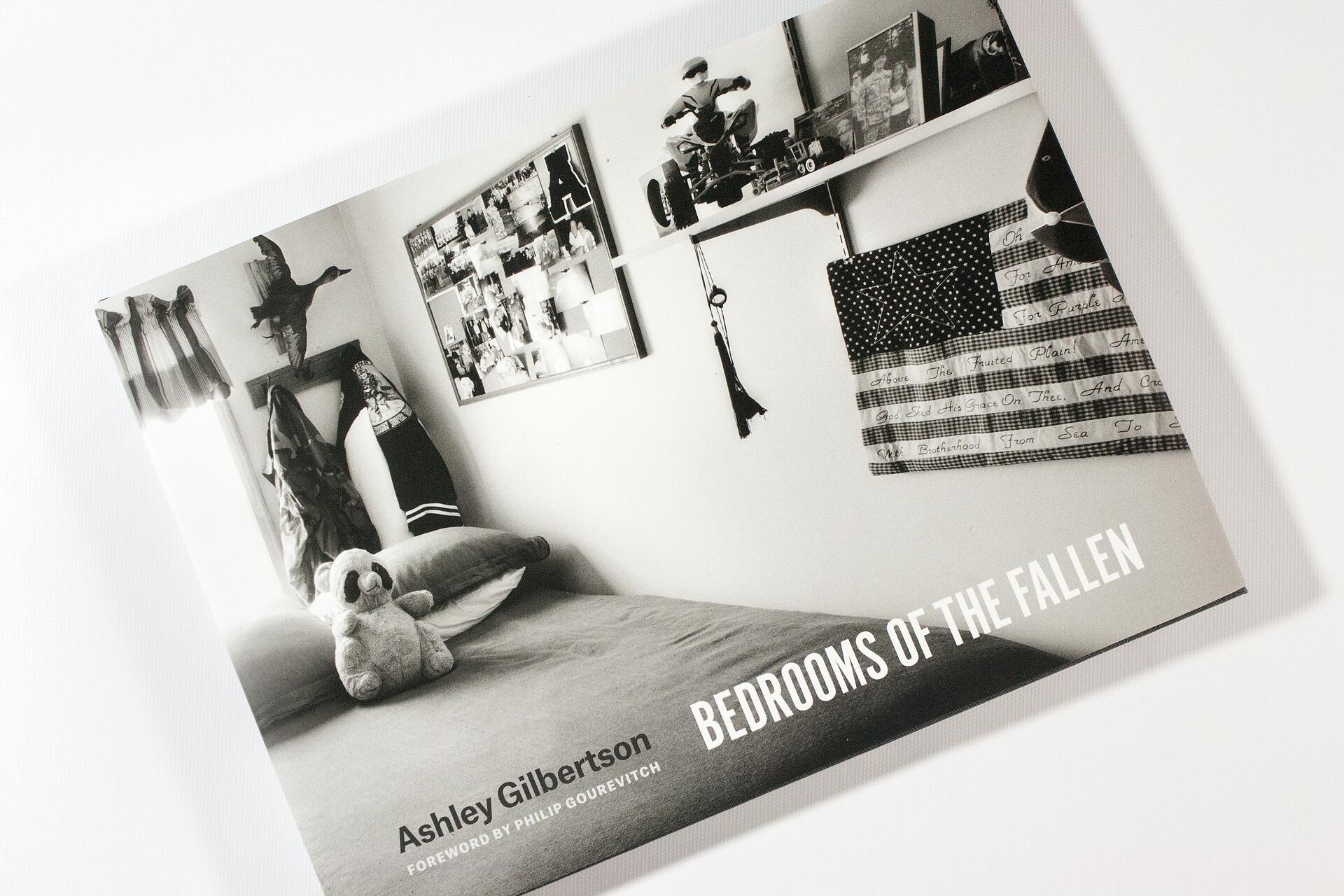 Black and white photograph of a bedroom with a made bed, a stuffed animal, photos on a bulletin board, and shelves with personal items. The image is the cover of a book titled "Bedrooms of the Fallen" by Ashley Gilbertson.