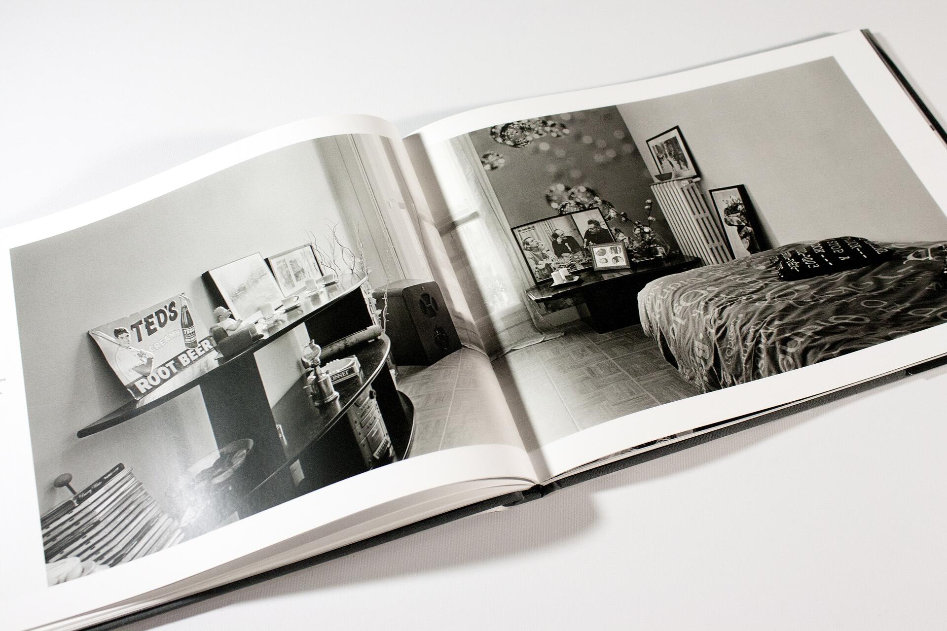 Open book showing a black and white photograph of a bedroom. The room features a bed with a patterned bedspread, a shelf with various items, framed pictures, and a window in the background.