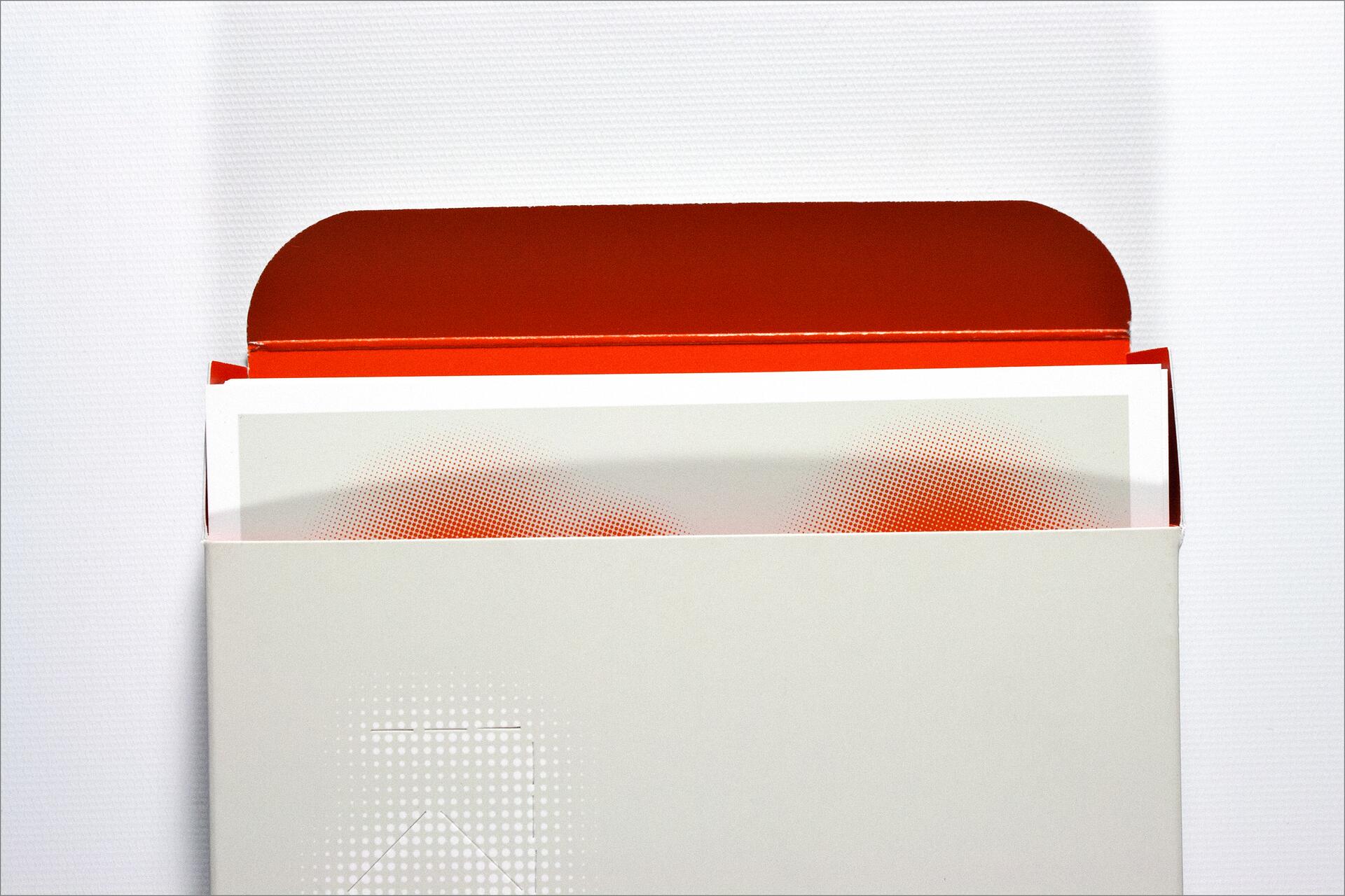 Open white box with a red inner flap revealing printed material inside, featuring red and white halftone patterns.