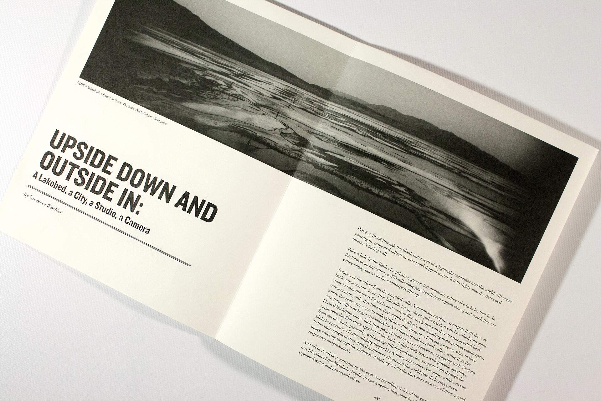 Open magazine spread showcasing a black and white photograph of a lakebed with surrounding hills. The headline reads, "Upside Down and Outside In: A Lakebed, a City, a Studio, a Camera." Text is displayed below the photograph.