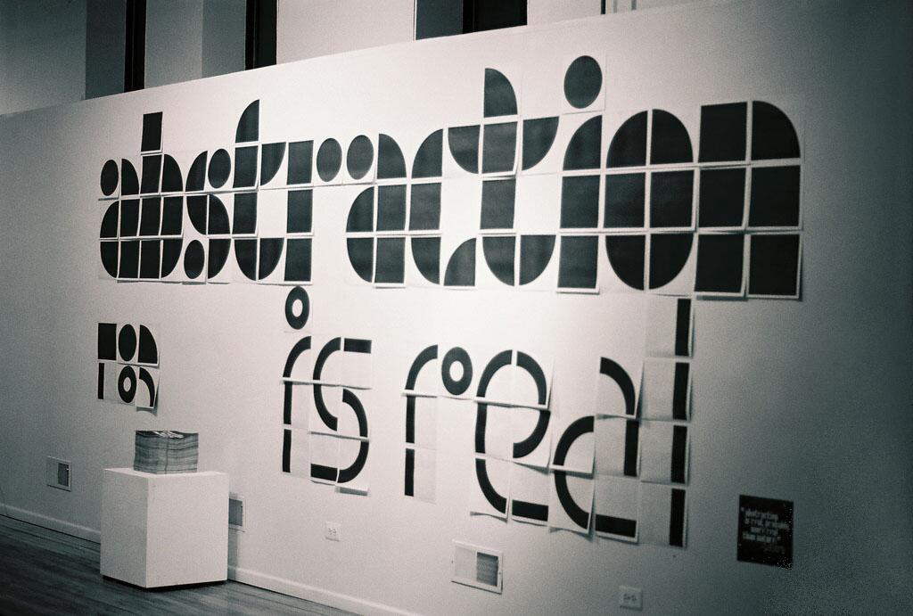 Abstract wall art installation with the phrase "abstraction is real" in stylized black letters on a white wall, composed of individual paper panels. Nearby, a small pedestal holds stacked paper. The setting is a gallery space with wooden floors.
