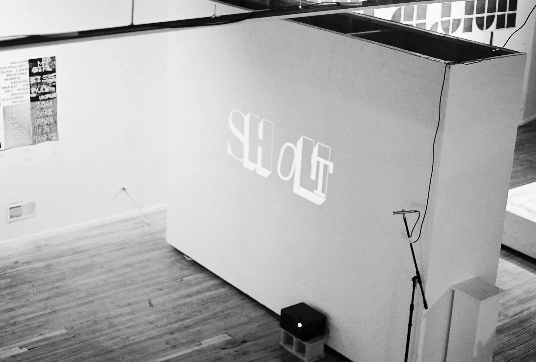 Black and white photograph of a gallery space with wooden floors. Wall text reading "SHOUT" is projected onto a white wall from a device on the floor. A microphone stand is nearby, and posters are displayed on the adjacent wall.