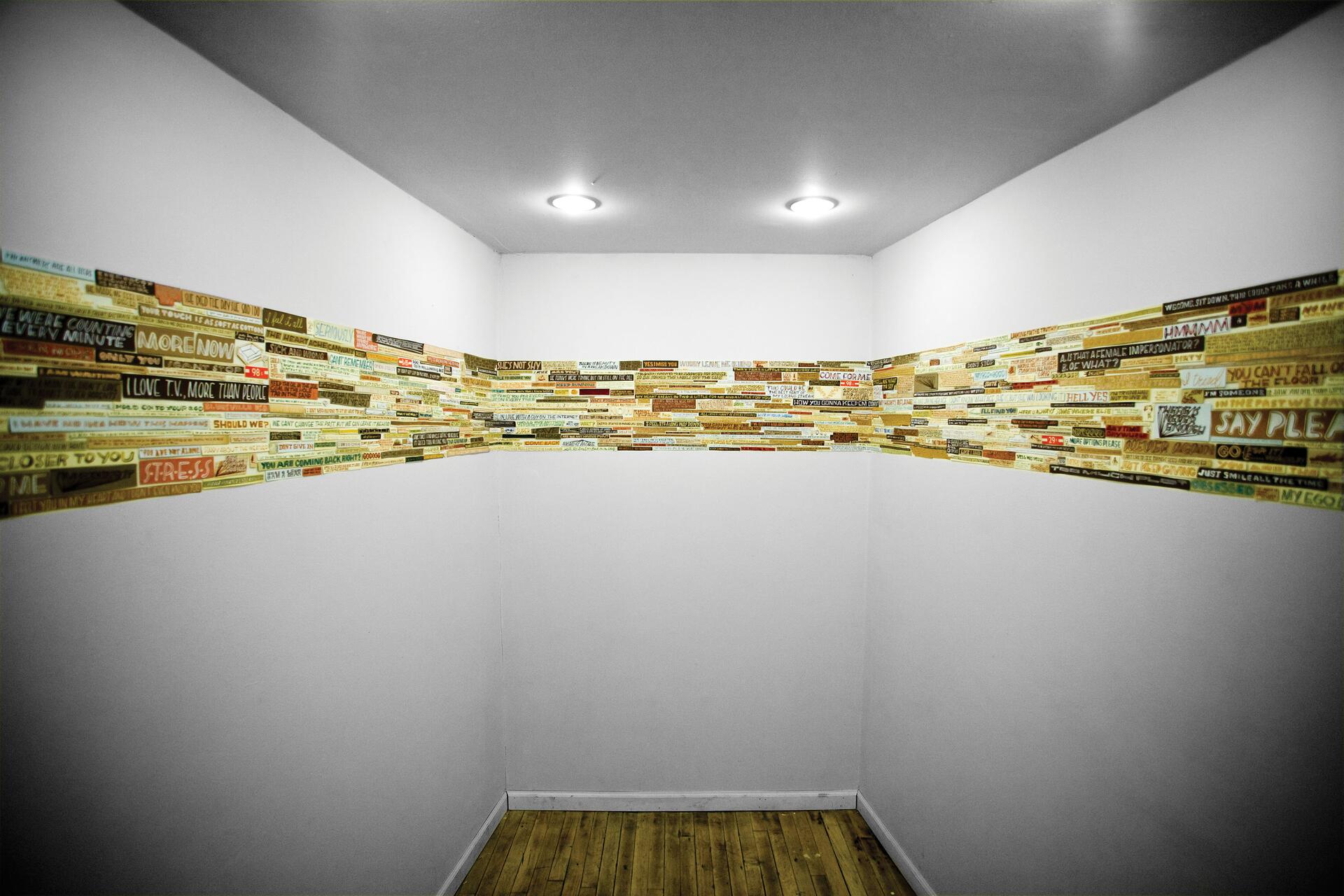 A corner of a room with wooden flooring features walls adorned with colorful text strips forming a collage around the perimeter. Two ceiling lights illuminate the minimalistic setting.
