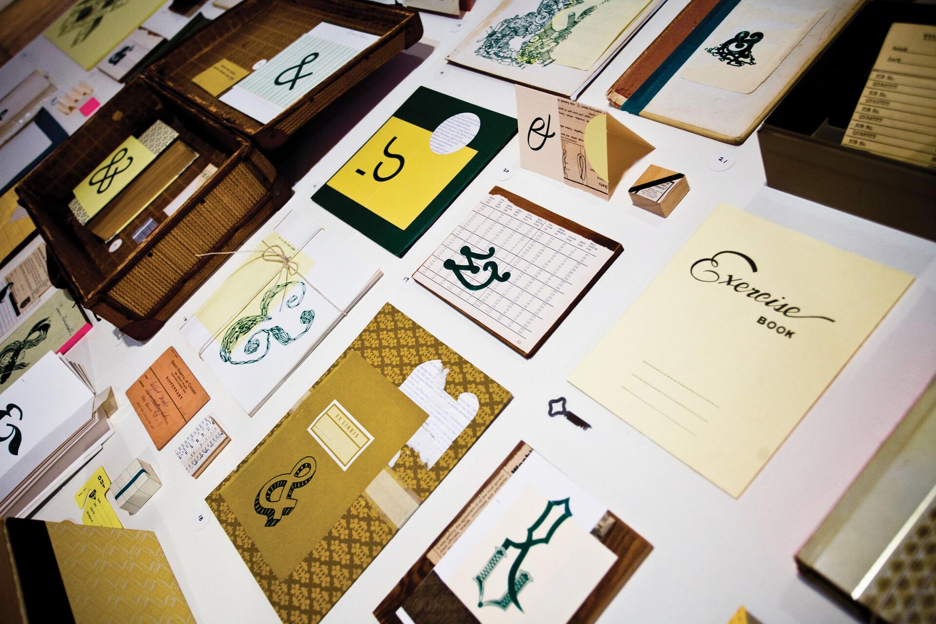 An assortment of vintage stationery items arranged on a table, featuring exercise books, envelopes, and cards, many with ornate typography and ampersand designs. A small suitcase is open on the left side.