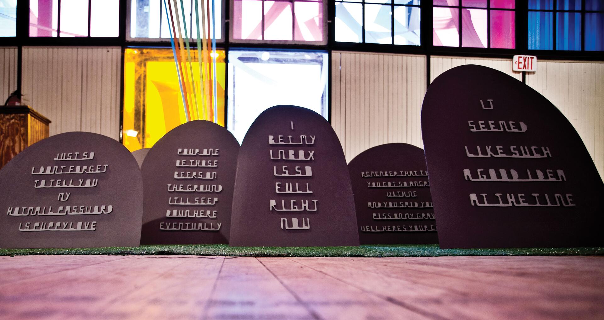 Art installation of tombstone-like structures with messages in cut-out letters, set in a colorful, illuminated indoor space with tall windows in the background.