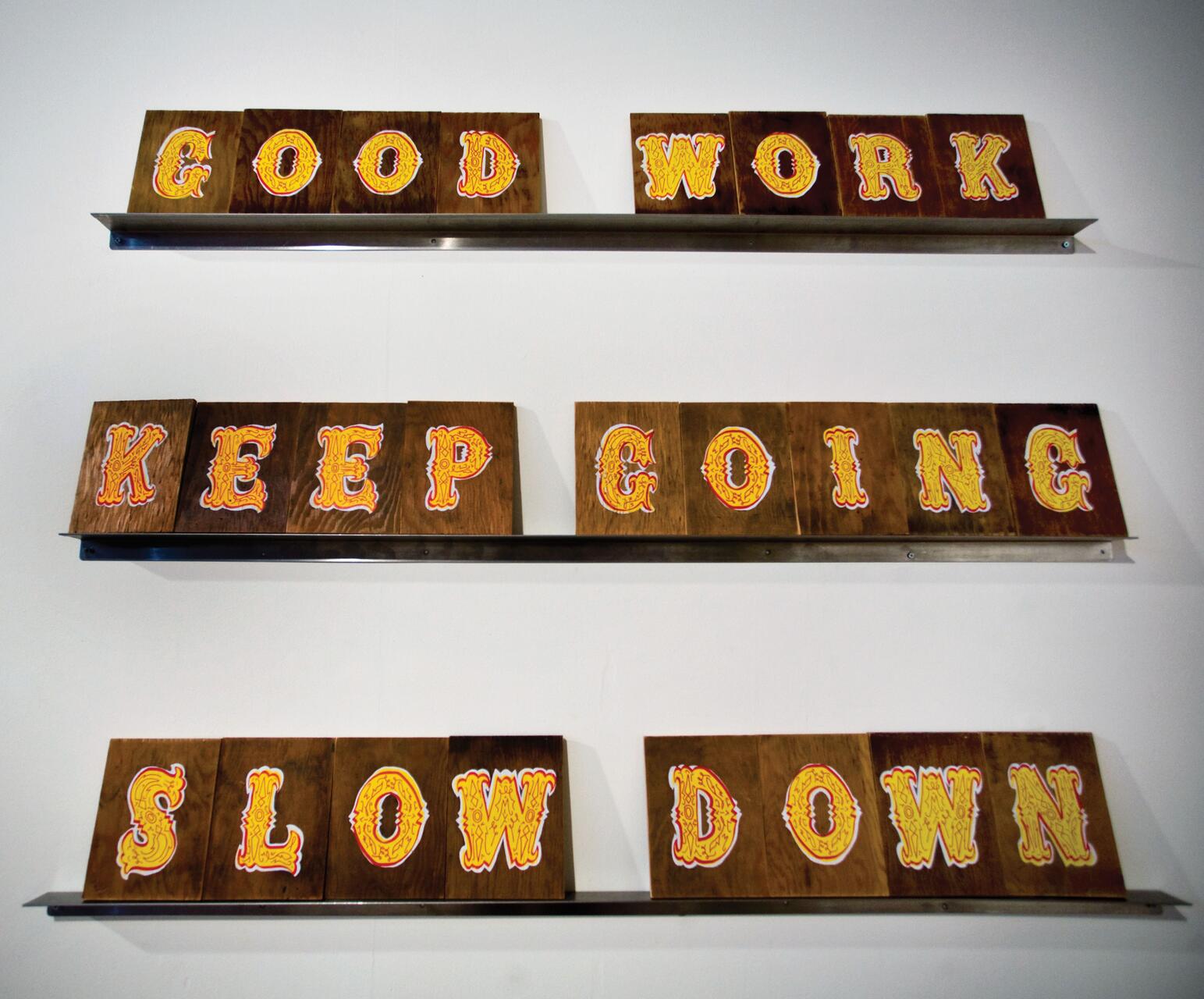 Wooden plaques on a wall displaying three phrases: "GOOD WORK," "KEEP GOING," and "SLOW DOWN," each in bright, decorative yellow and orange letters on a dark wood background.