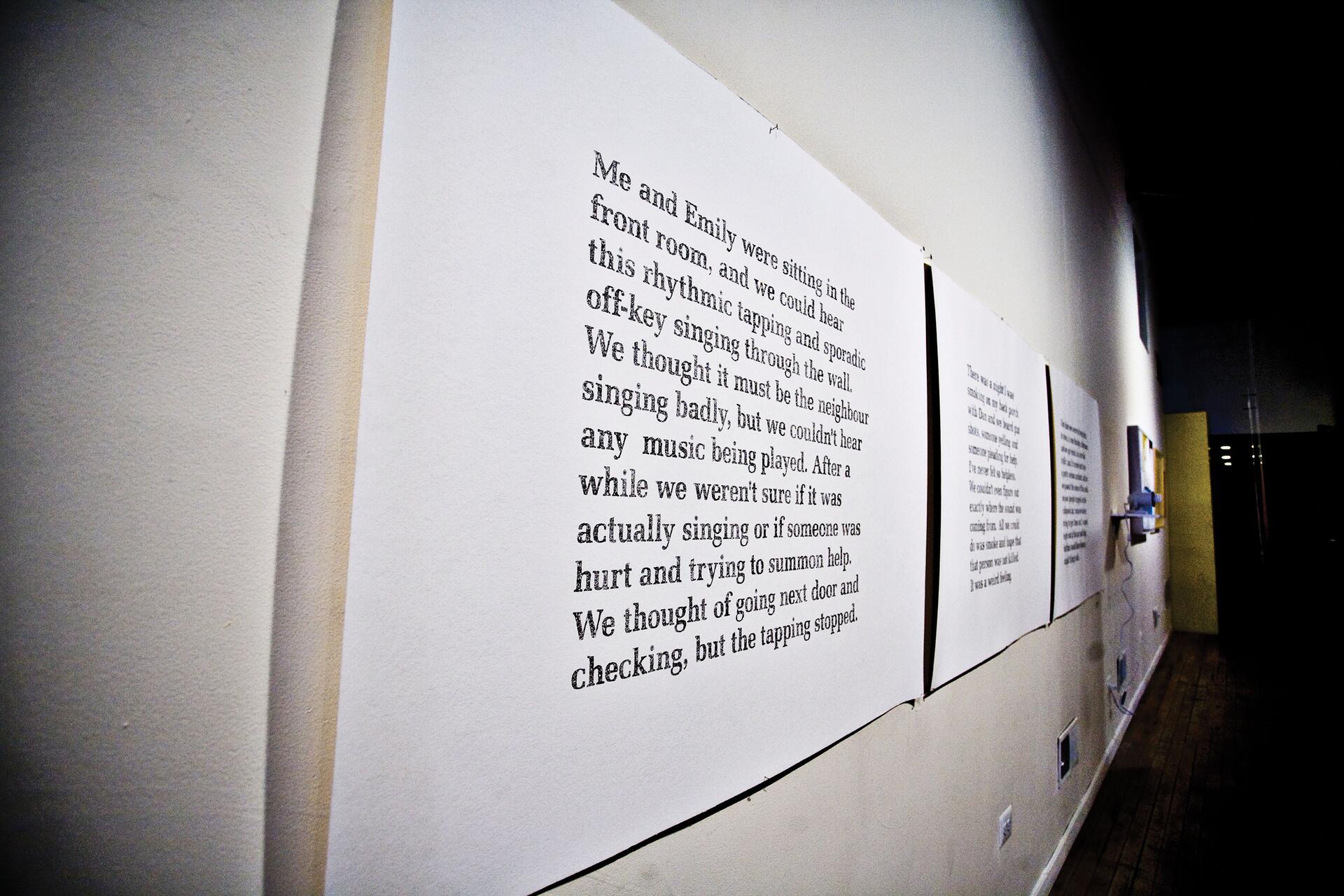 Large sheets of paper with typed text mounted on a gallery wall, extending down a corridor. The text is printed in a serif font. The setting is a dimly lit space with wooden flooring.