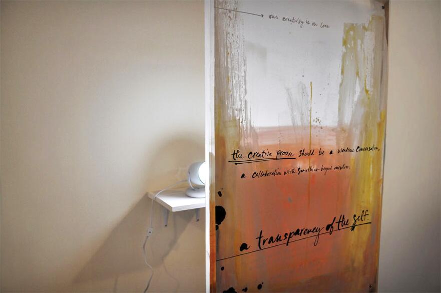 A dimly lit room with a wall art piece featuring abstract orange and yellow paint with handwritten text. A small white shelf holds a round light, casting a shadow on the beige wall.