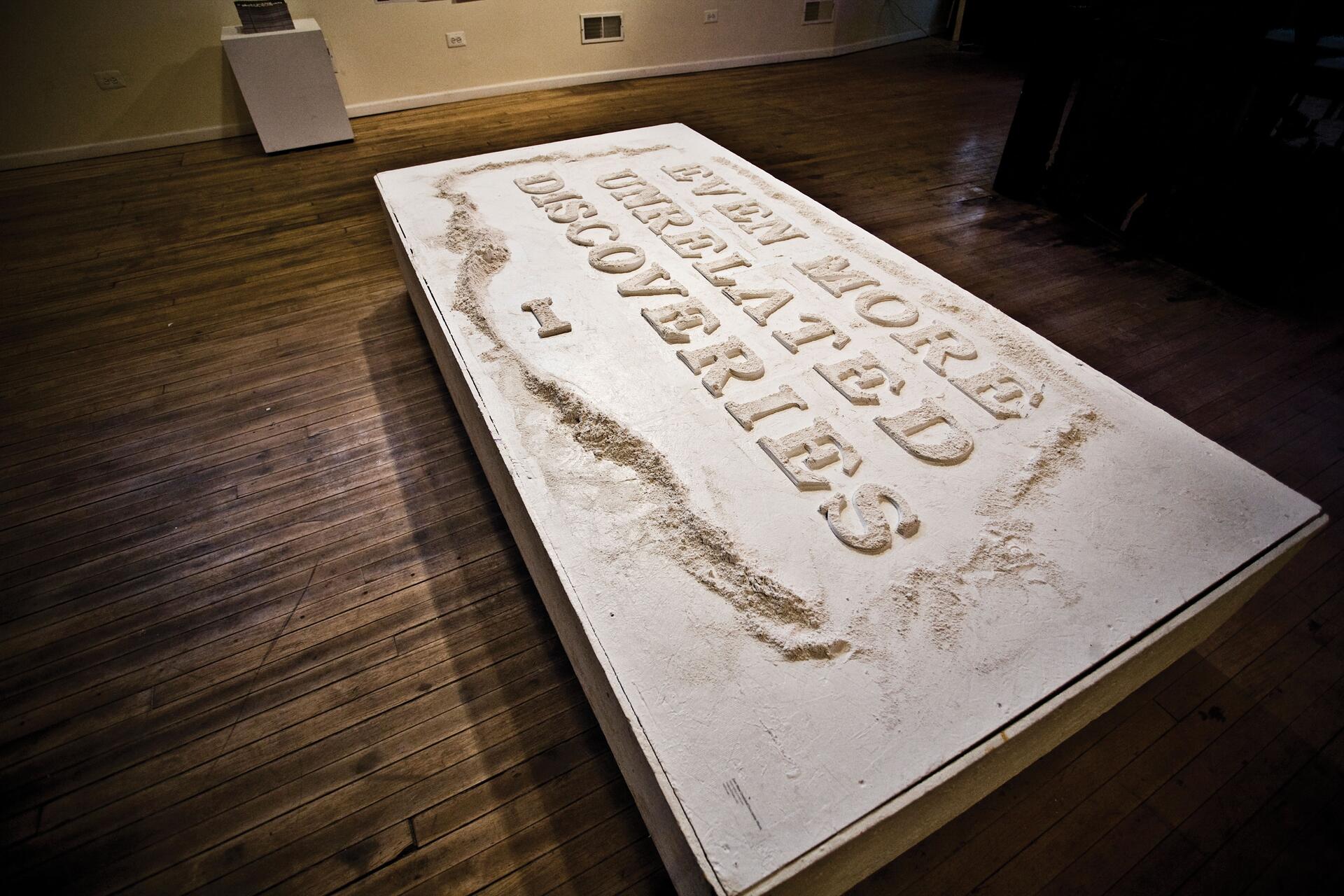 Large white sculpture resembling a book cover with embossed text on a wooden floor in a gallery setting.