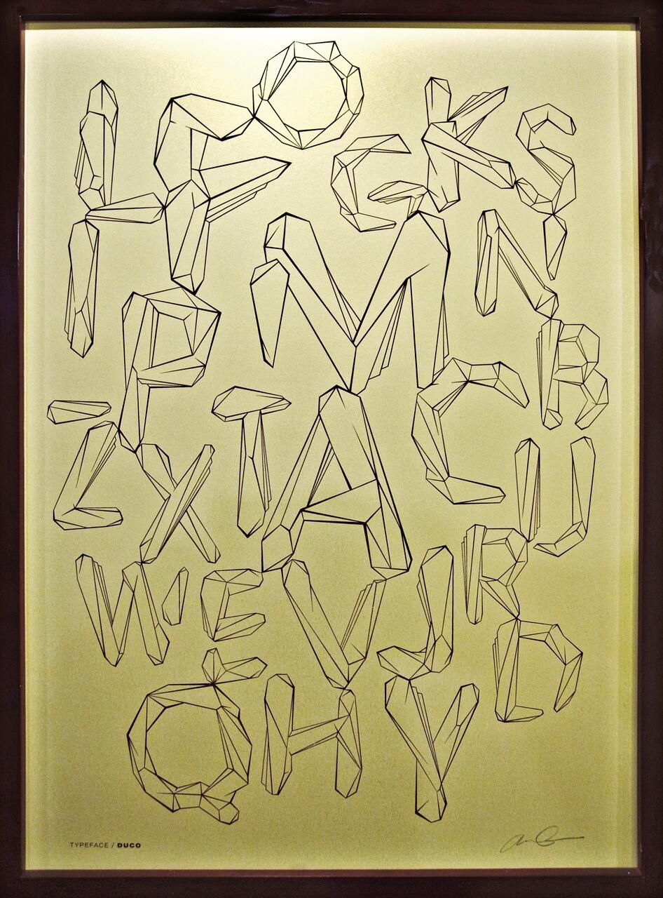 Geometric lettering artwork with angular, faceted letters in black on a light yellow background, framed in dark wood.