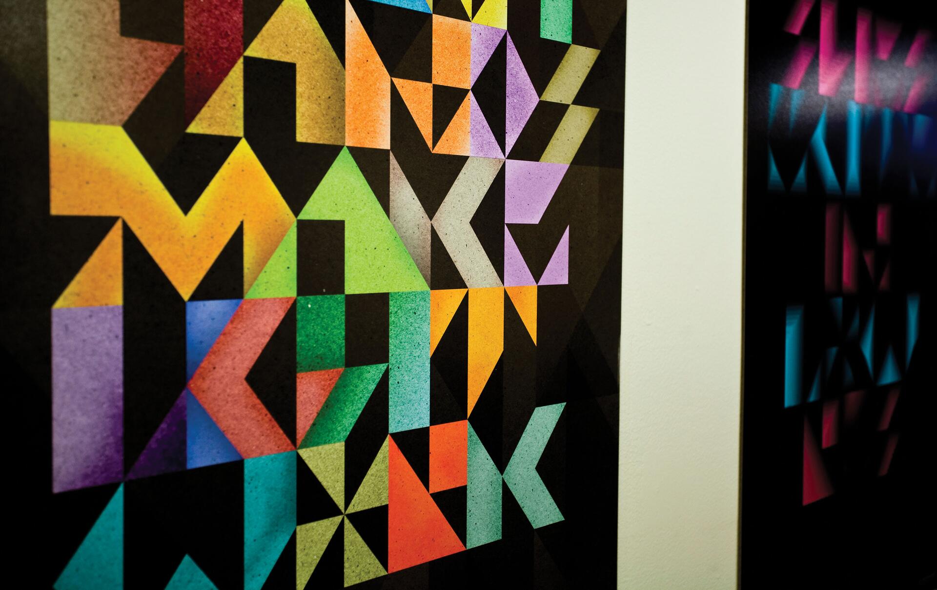 Geometric art poster with colorful triangular patterns in shades of orange, green, purple, and blue on a black background. Another poster with similar patterns, but brighter shades, is partially visible to the right.