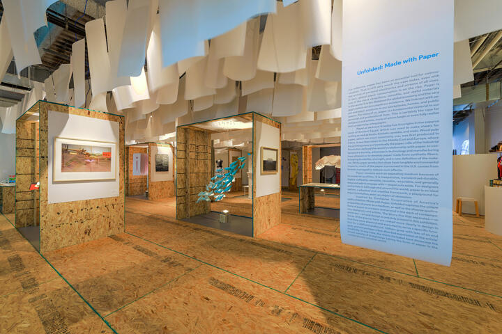Art gallery installation with hanging paper structures, wooden panels displaying framed artwork, and an informational panel on the right. The atmosphere is creative and contemporary.