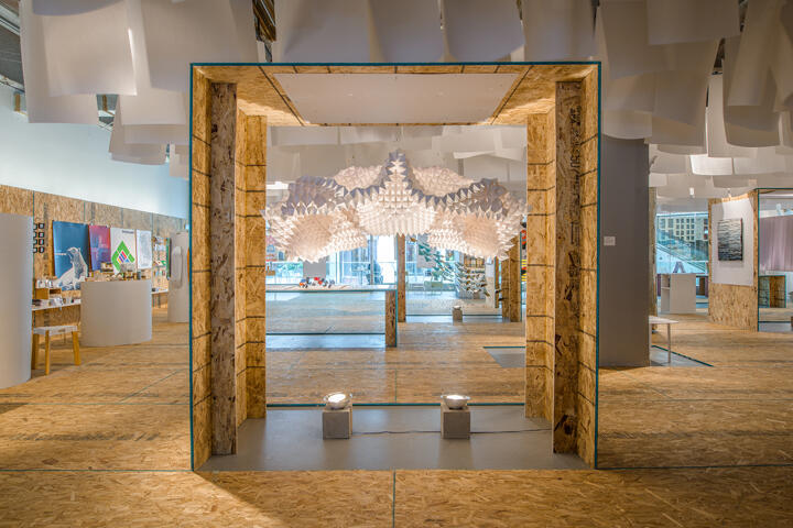 Indoor art exhibition room with wooden and white decorations. A geometric paper sculpture hangs from the ceiling, surrounded by plywood walls and floor. Various artworks are displayed on the perimeter, with soft, ambient lighting enhancing the modern aesthetic.