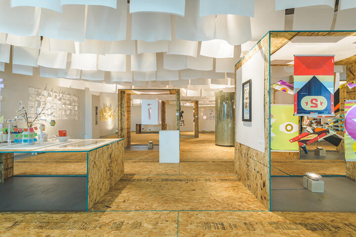 Modern art gallery with white, suspended ceiling panels and OSB wood displays. Colorful geometric art pieces and framed artworks are exhibited. The space has a bright, minimalist aesthetic.