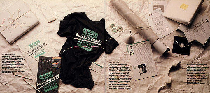 A black t-shirt with green text lies on crumpled paper, surrounded by brochures, a pencil, wrapped parcels, a newspaper, and a spool of twine.