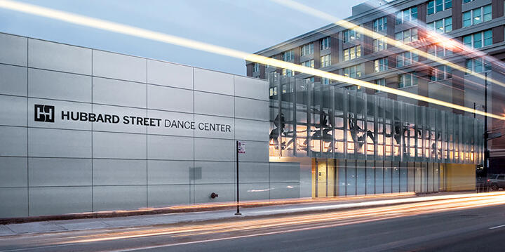 Hubbard Street Dance Center building exterior with modern design, featuring large glass windows and a sleek facade. A long exposure captures streaks of light from passing traffic on the adjacent street.
