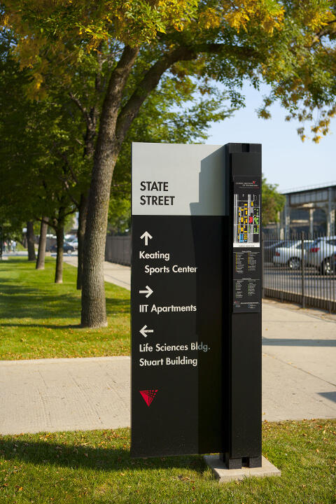 Directional sign with arrows pointing to Keating Sports Center, IIT Apartments, Life Sciences Building, and Stuart Building, set against a backdrop of a tree-lined street and sidewalk in sunlight.