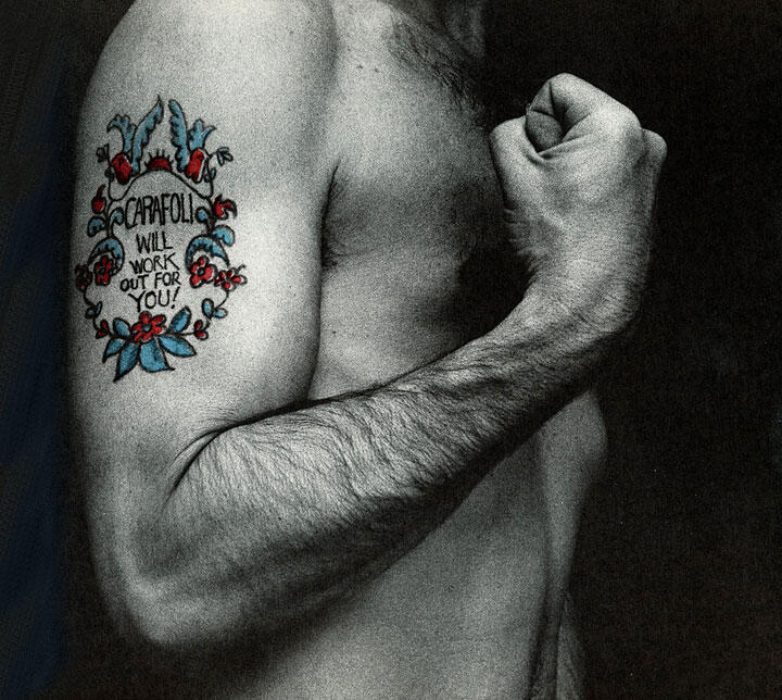 Tattooed arm with colorful design, featuring red and blue flowers and birds. The person is clenching their fist, showcasing the tattoo on their bicep. The background is dark, highlighting the tattoo's details and colors.