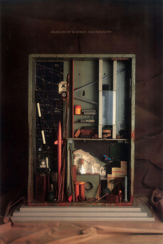 A shadow box with scientific instruments, a rocket model, constellations map, and various small objects, set against a dark background, framed by draped fabric.