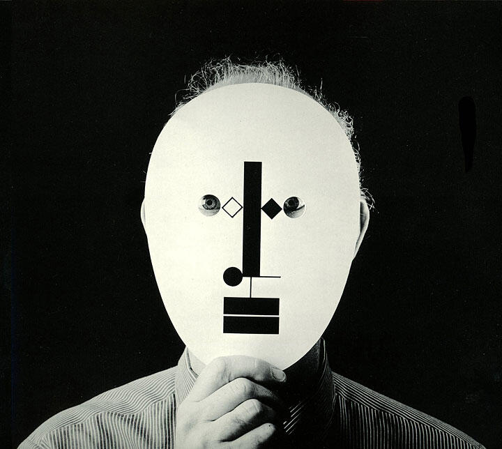 Person holding a mask with abstract geometric patterns resembling a face, captured in black and white against a dark background.