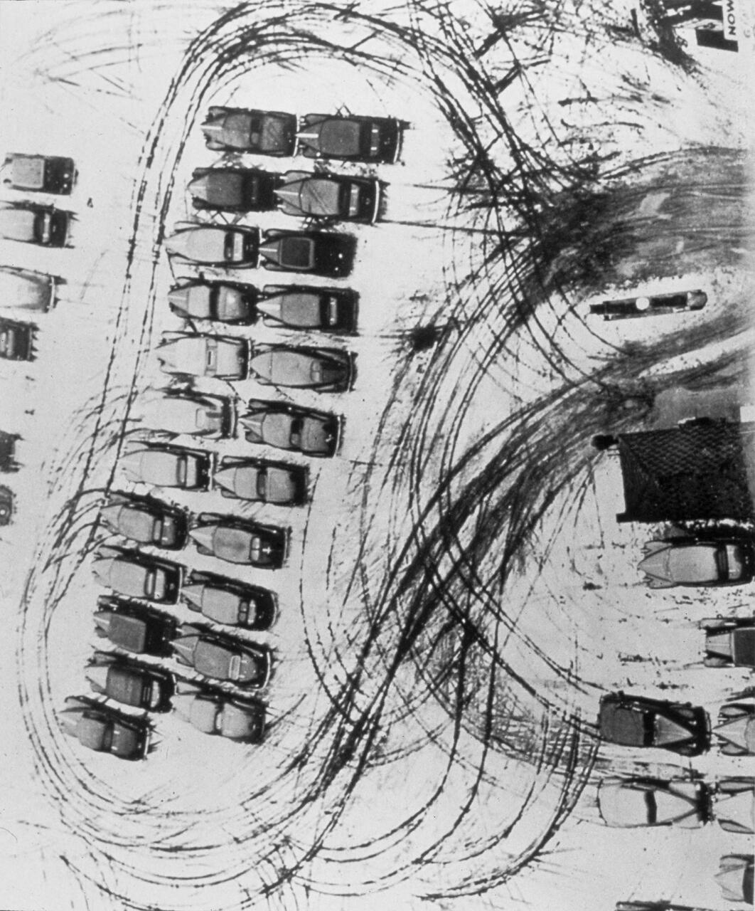Aerial view of numerous cars parked in rows on a snowy surface with chaotic tire tracks creating sweeping curves across the scene, suggesting movement and activity.