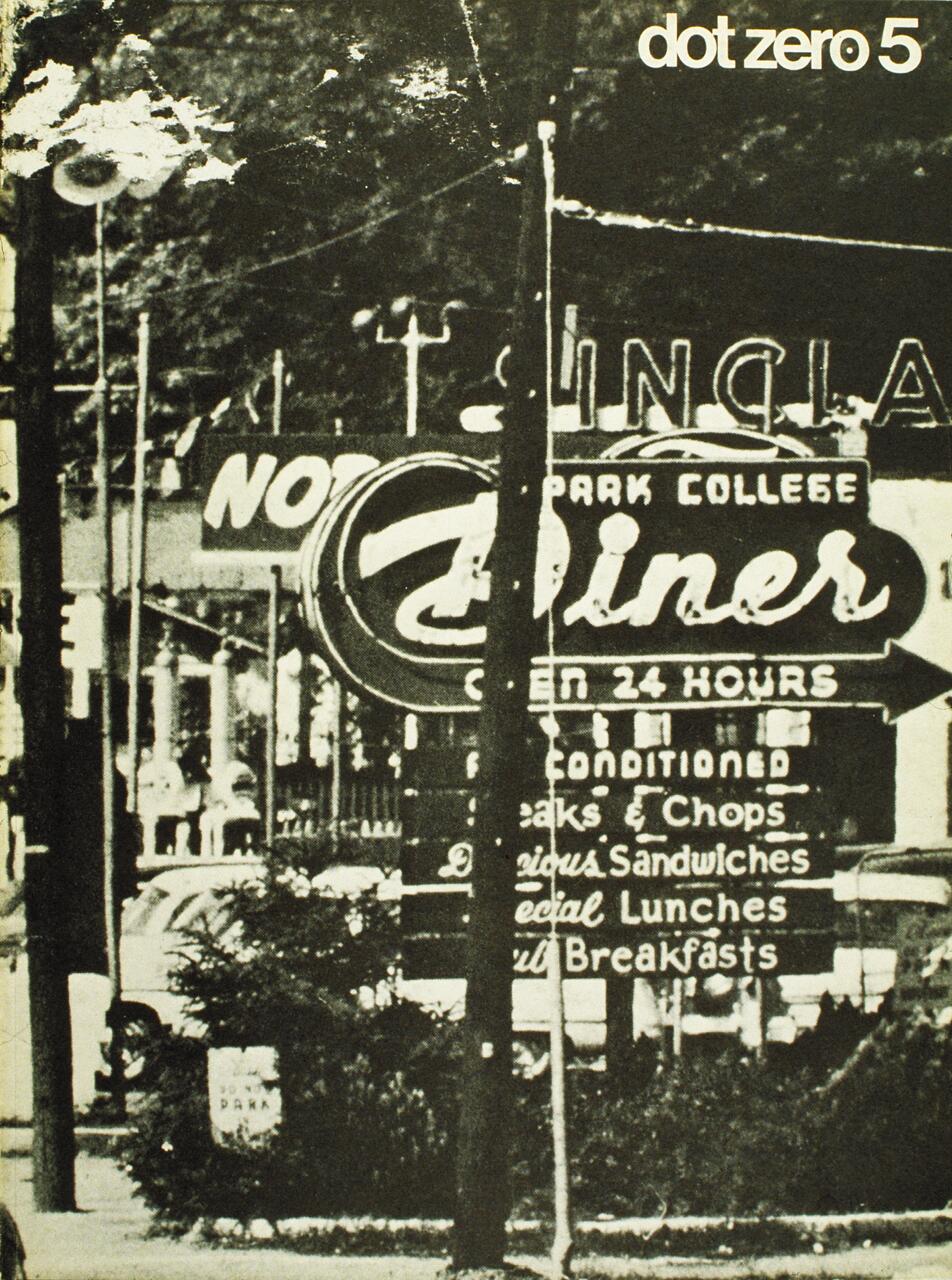 Black-and-white photograph of a retro diner sign reading "Open 24 Hours" and advertising various meals. The background shows blurred elements of a street and vintage cars.