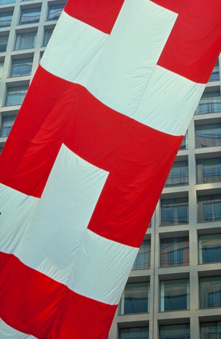 Large Swiss flag with a white cross on a red background, hanging in front of a multi-story building with visible windows.