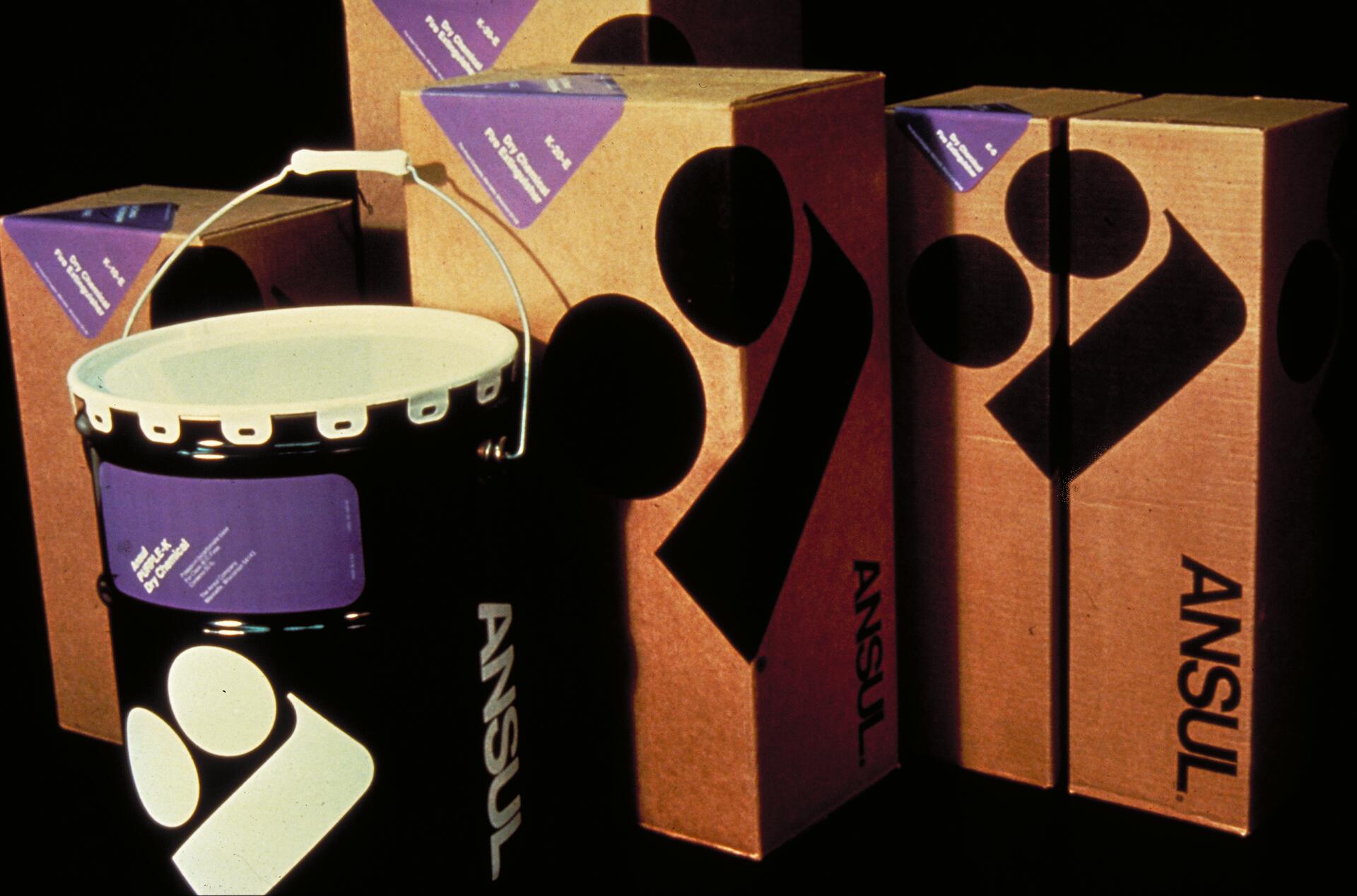 Containers and boxes with the ANSUL logo, featuring large black graphics and purple labels, set against a dark background.