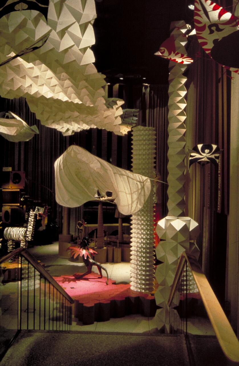 Surreal indoor installation with geometric shapes and paper-like structures lit dramatically. An abstract figure with colorful wings is in the center, surrounded by hanging and standing sculptures, creating an artistic and whimsical atmosphere.