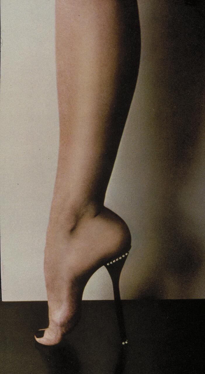 A person balancing on tiptoe, foot positioned to mimic the arch of a high-heeled shoe, with a stiletto heel attached to the heel. The skin appears smooth, with neutral tones against a plain background.