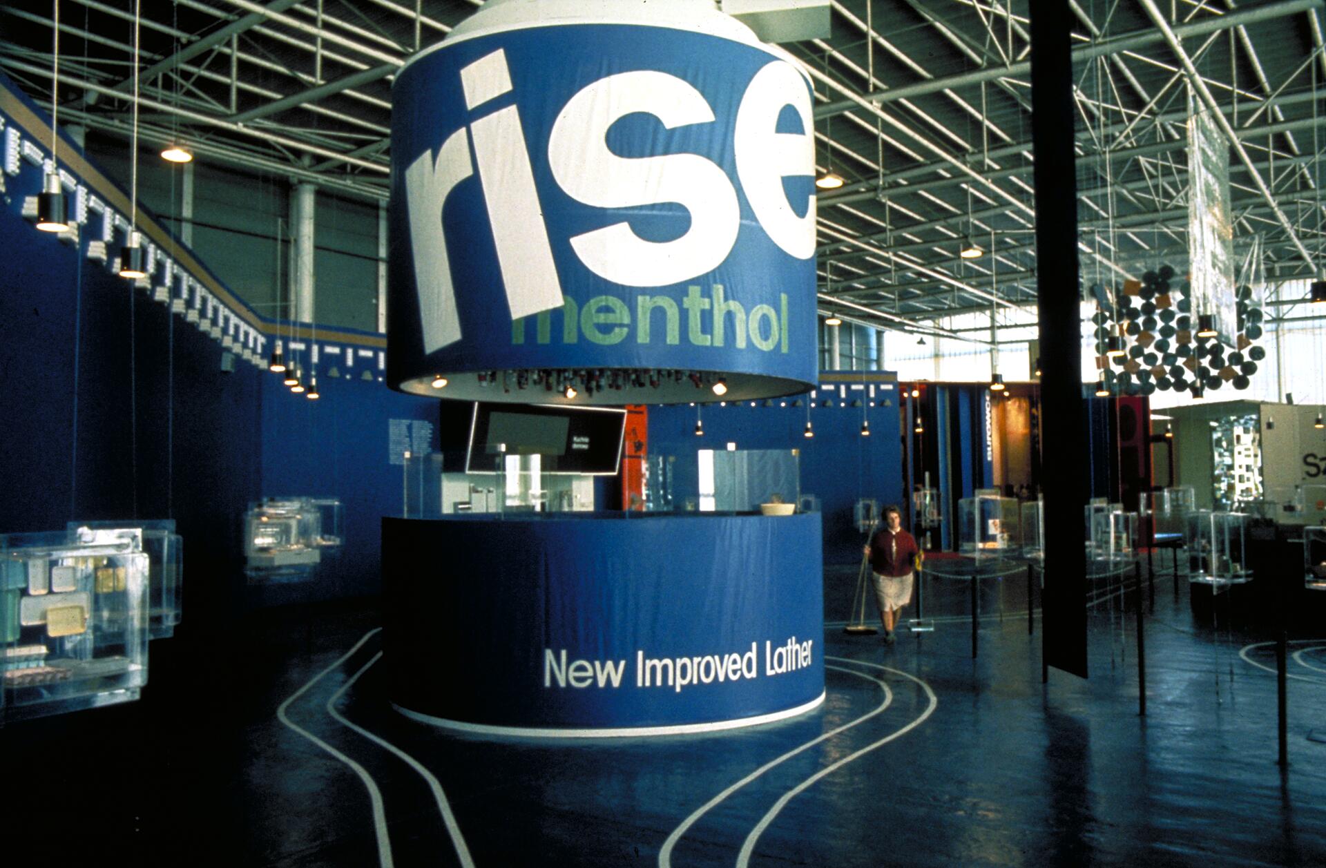 Exhibition booth with large blue cylindrical display featuring the word "rise" and "menthol," surrounded by modern exhibits and a person walking on a polished blue floor inside a spacious industrial hall with exposed metal beams.