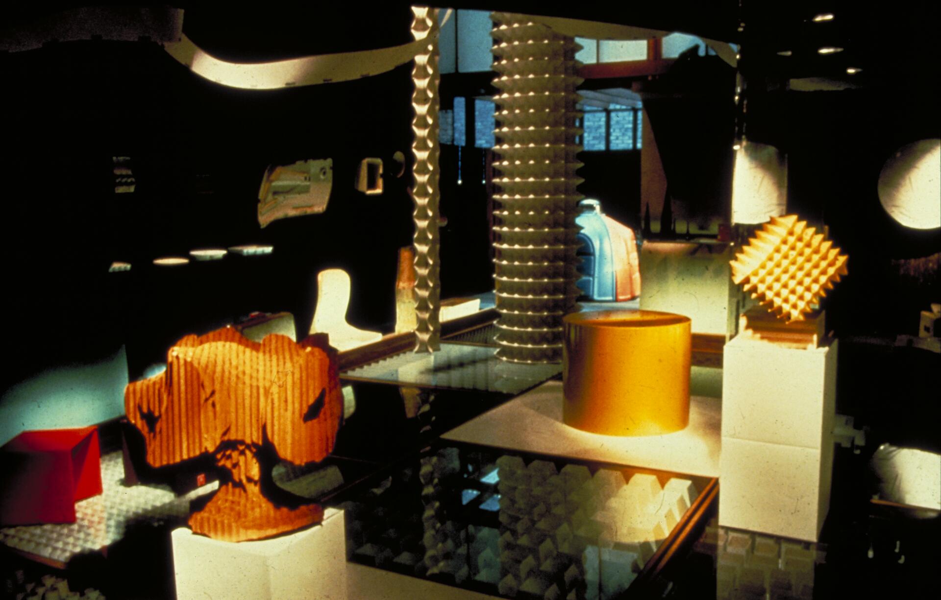 Exhibition display of abstract sculptures on pedestals, featuring a wooden textured piece, a yellow geometric shape, and other colorful forms. The setting is dimly lit with a modern, industrial atmosphere.