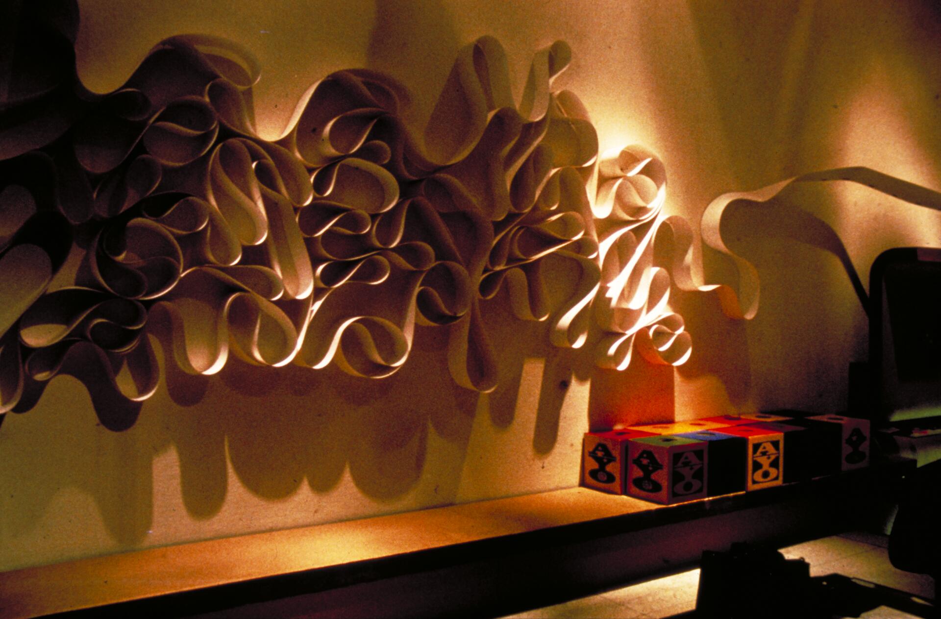 Abstract paper sculpture with intricate loops casting dramatic shadows on a wall, alongside colorful geometric cubes on a shelf, illuminated by warm lighting.