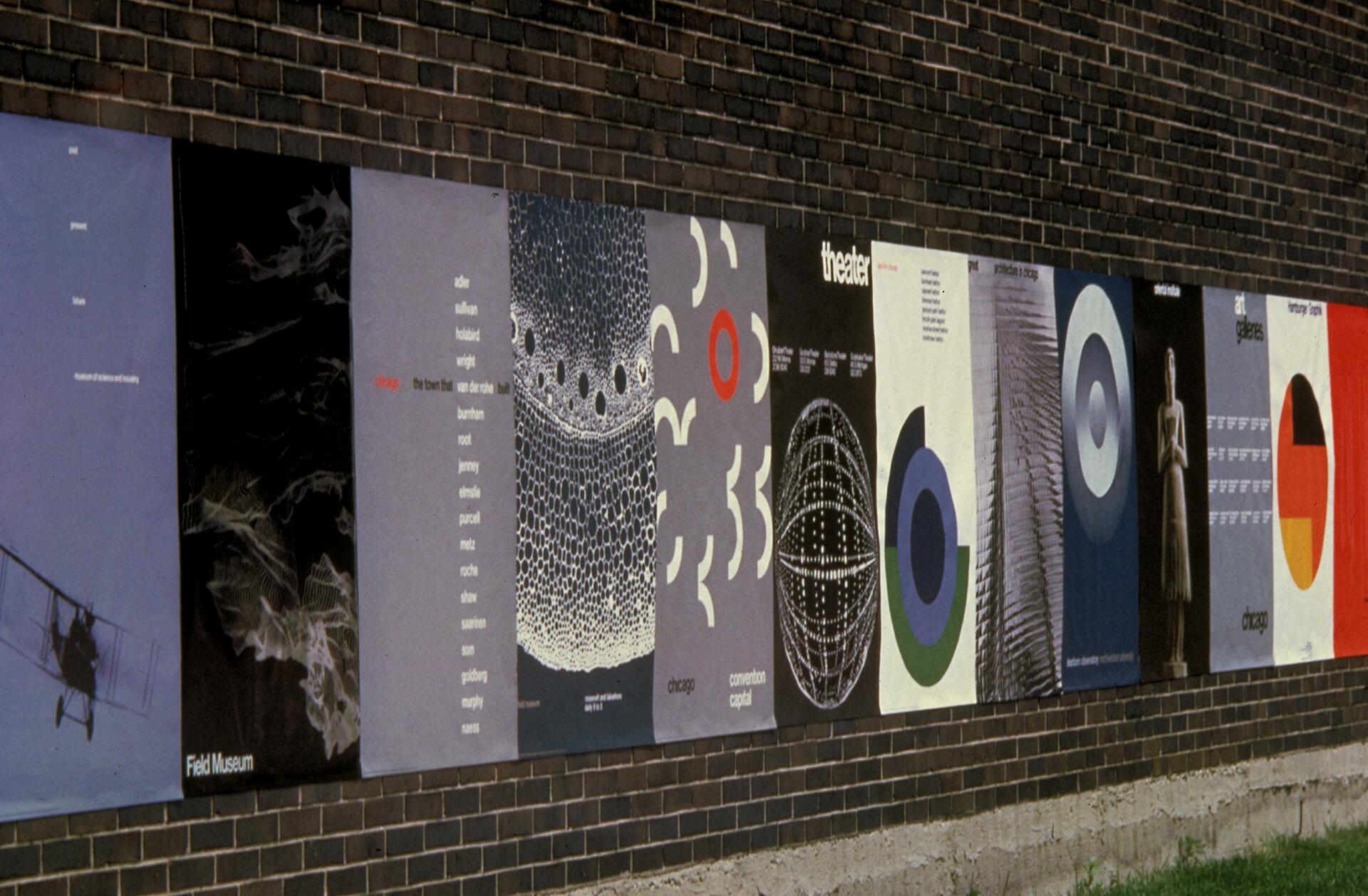 A row of colorful posters displayed on a brick wall, featuring various abstract and geometric designs, including circles, patterns, and architectural elements, each conveying a distinct artistic style.