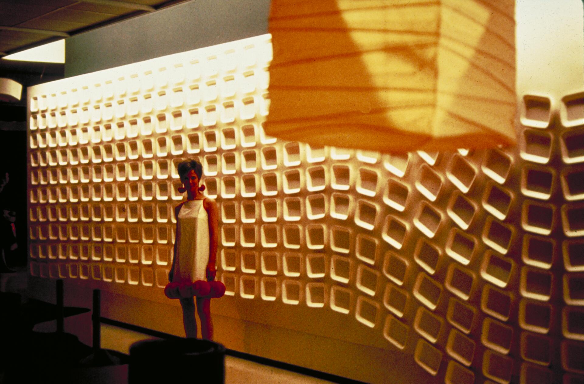 Woman in a white dress stands in front of a wall with a pattern of recessed squares, illuminated by warm lighting. A large, abstract orange lampshade hangs in the foreground.