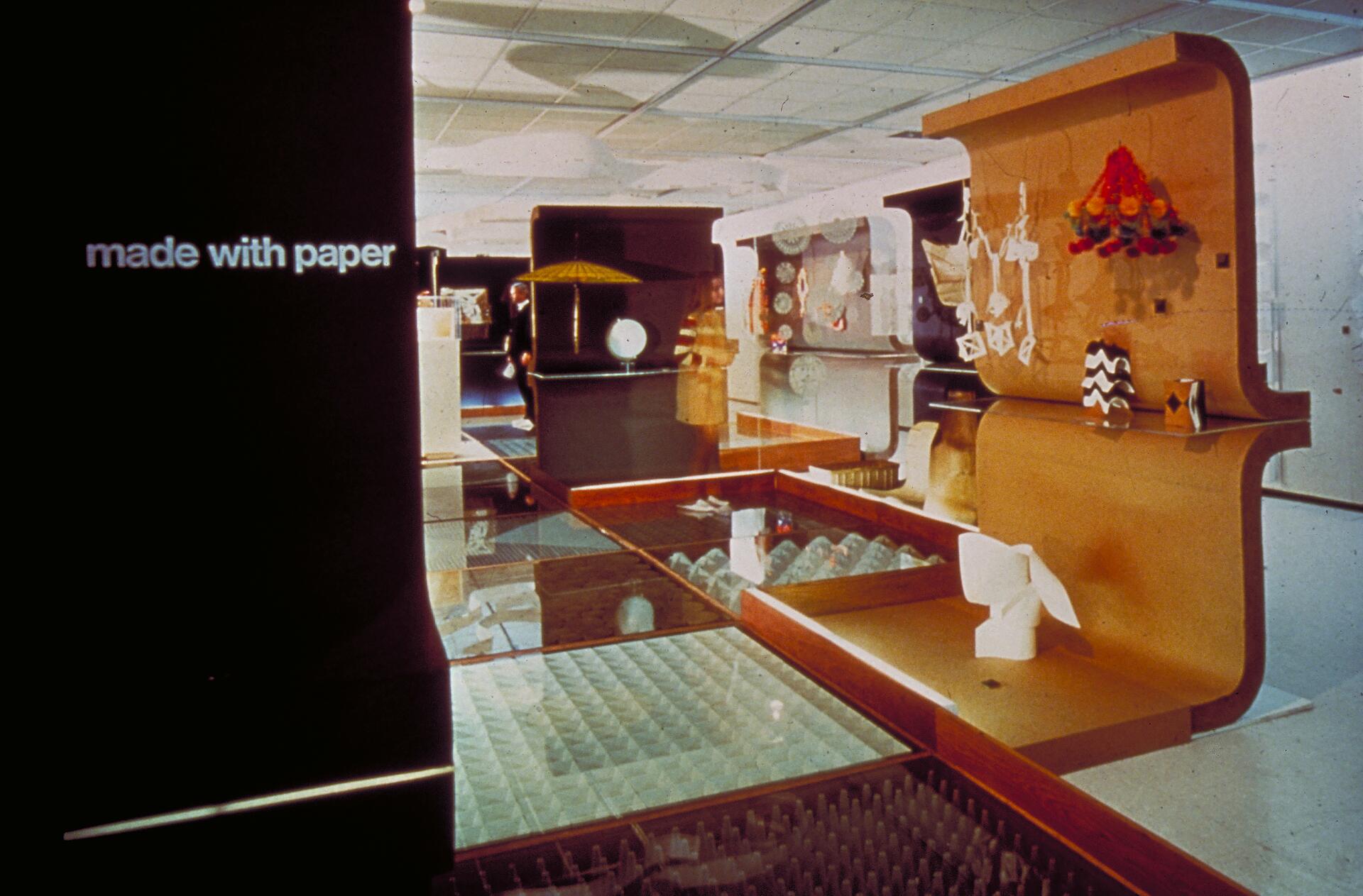 Exhibit showcasing various paper crafts and sculptures, displayed against a backdrop of curved, wooden panels. A soft, warm glow illuminates the scene, highlighting models and paper art.