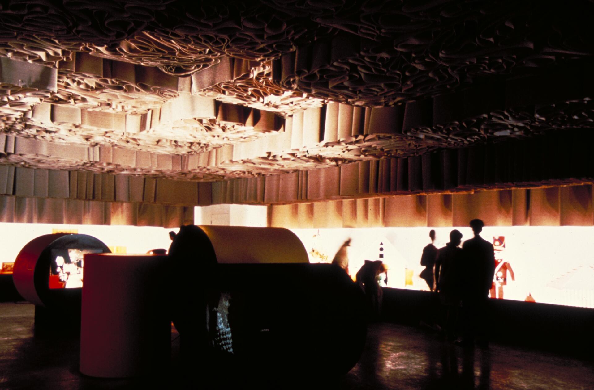 Dimly lit room with an intricate, textured ceiling and large cylindrical structures on the floor. Silhouetted people stand in front of a brightly lit wall with colorful displays, creating a contrast with the dark surroundings.