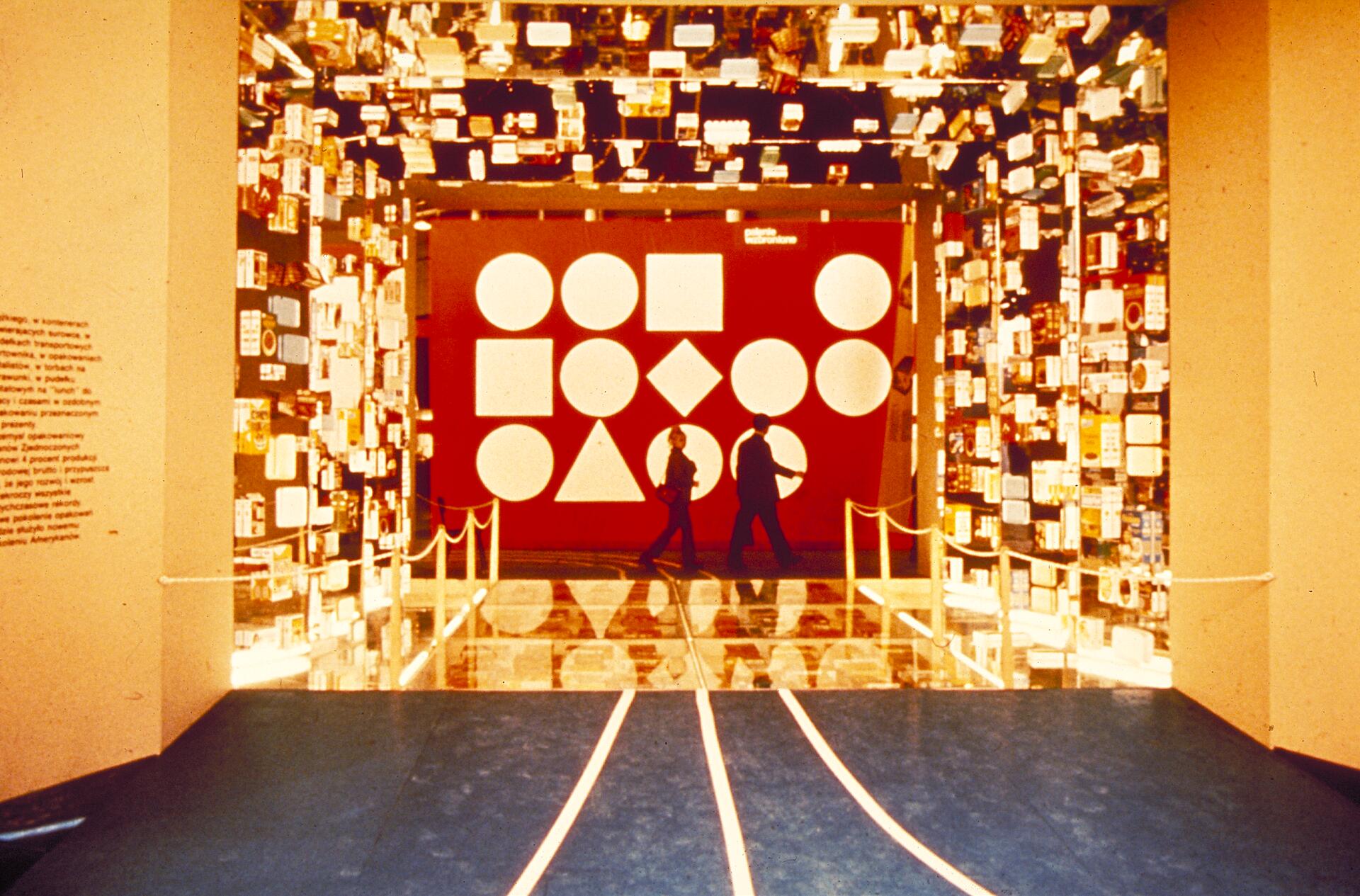A hallway with reflective walls displaying various logos leads to a red wall with geometric shapes, including circles and squares. Two people walk on a glossy floor with white rope barriers.