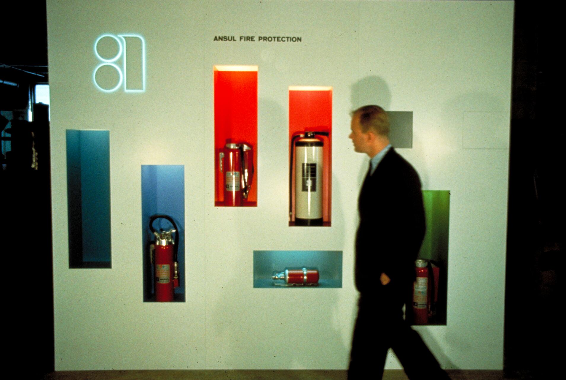 Man in a suit walks past a white display wall with recessed shelves holding various fire extinguishers in red, blue, and green accents.