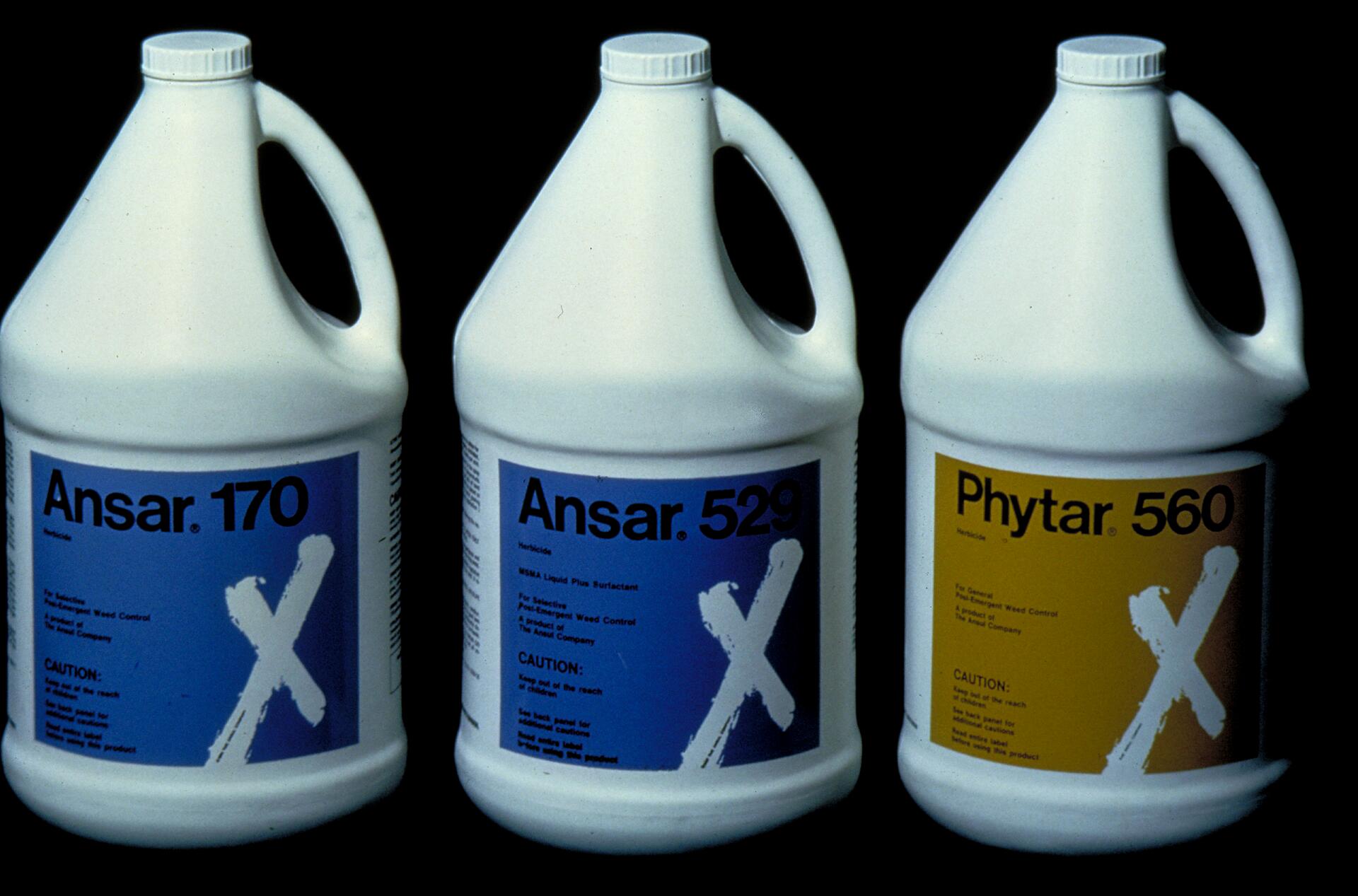 Three white gallon containers with handles, labeled "Ansar 170," "Ansar 529," and "Phytar 560." Labels have blue and yellow backgrounds with a large white "X." Containers are arranged side by side against a black background.