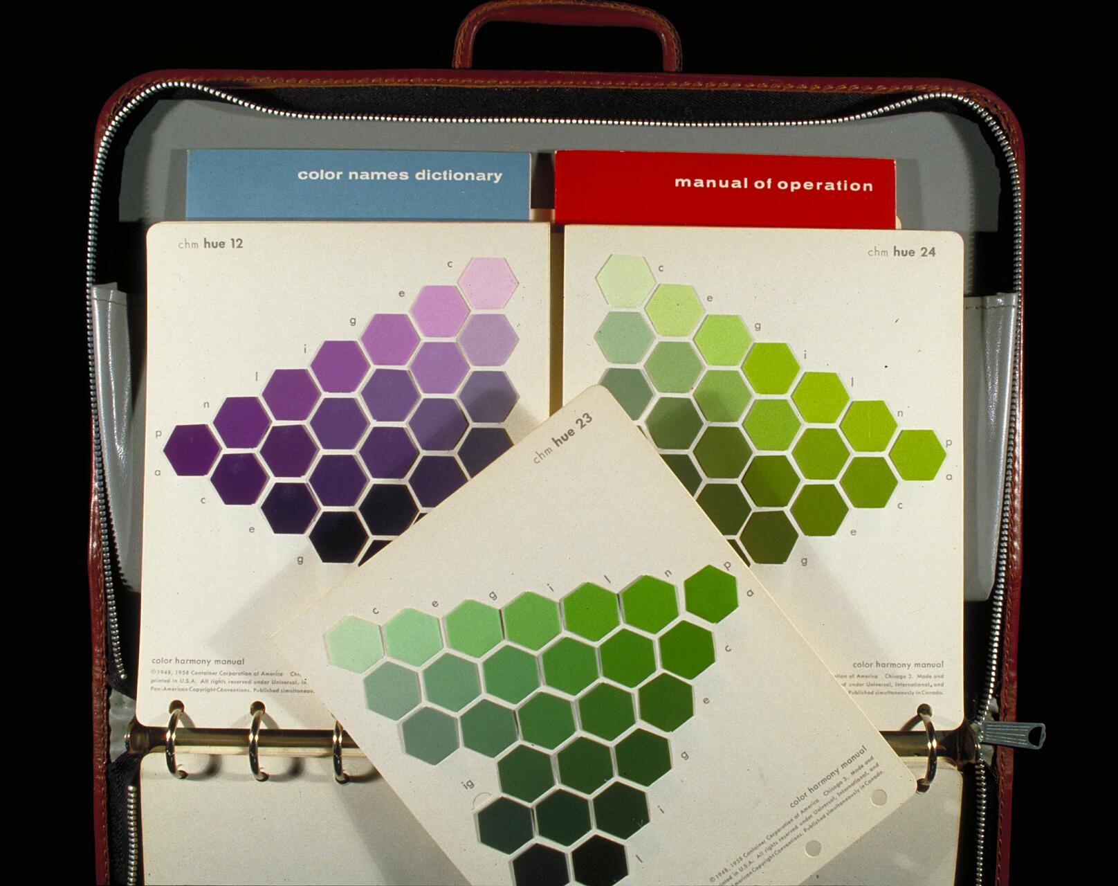 Open binder with color swatches in hexagon patterns, showing shades of purple and green. Two manuals in blue and red, titled "color names dictionary" and "manual of operation," are visible in the binder pockets.