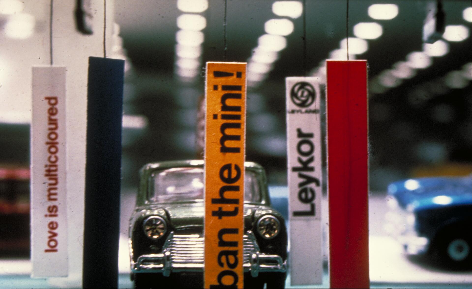 Small classic car model partly obscured by colorful vertical banners with text, set in a bright showroom with blurred lights in the background.