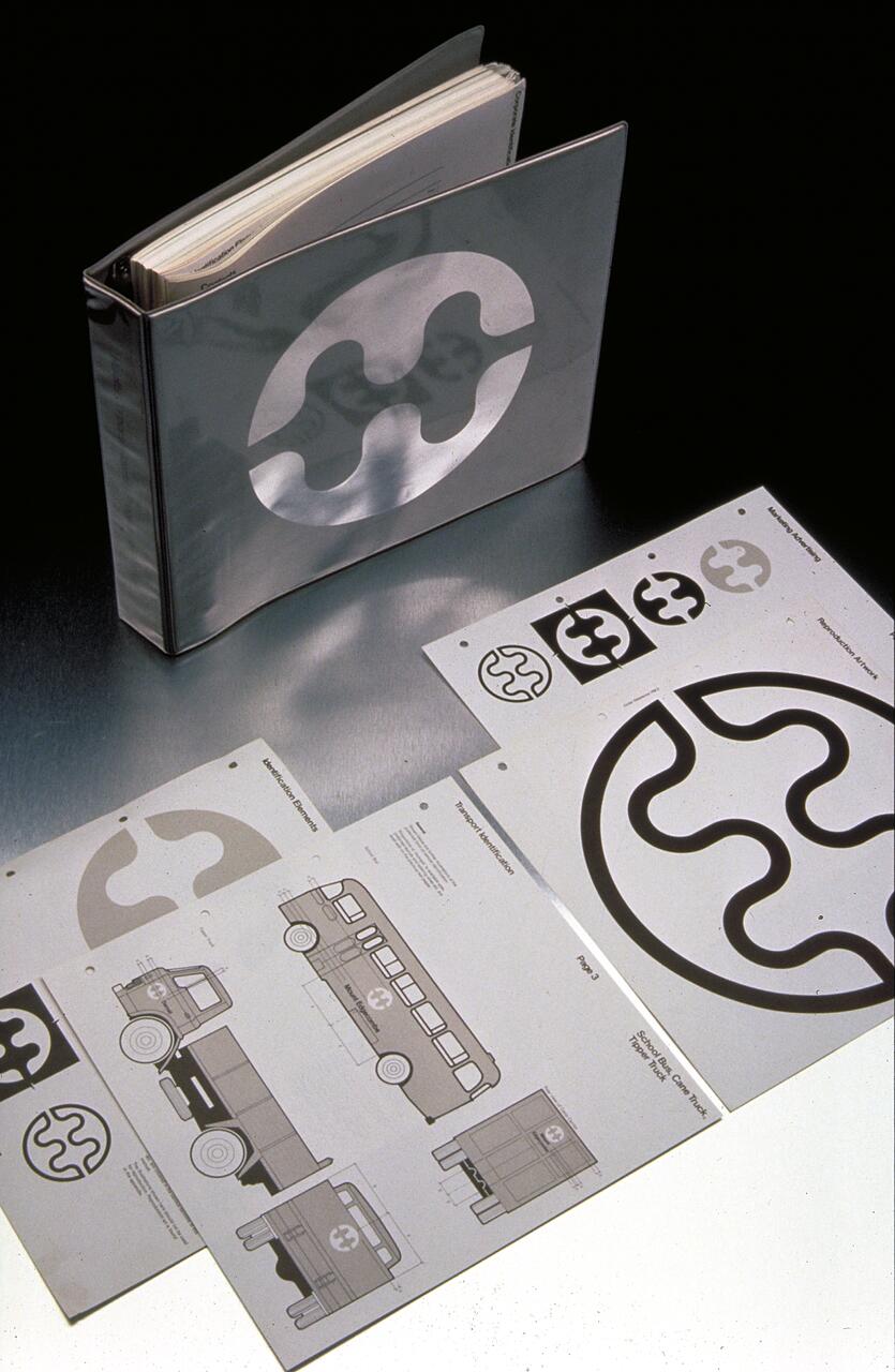 A binder with a metallic cover featuring a circular abstract logo, surrounded by graphic design sheets displaying vehicle illustrations and logo variations on a dark surface.