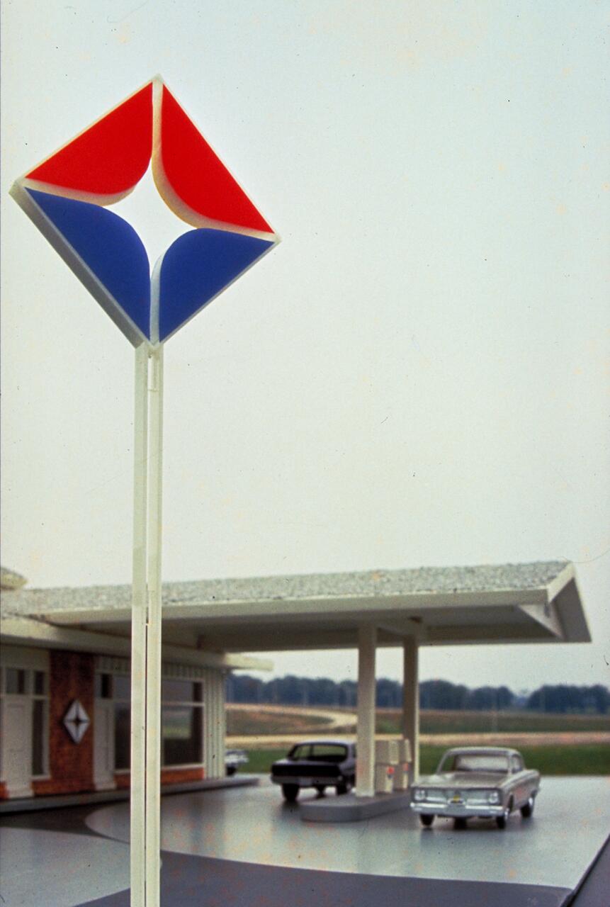 A retro gas station model with a red and blue star emblem on a tall sign. The station features two vintage cars parked under a canopy, with a building in the background and an open field beyond.