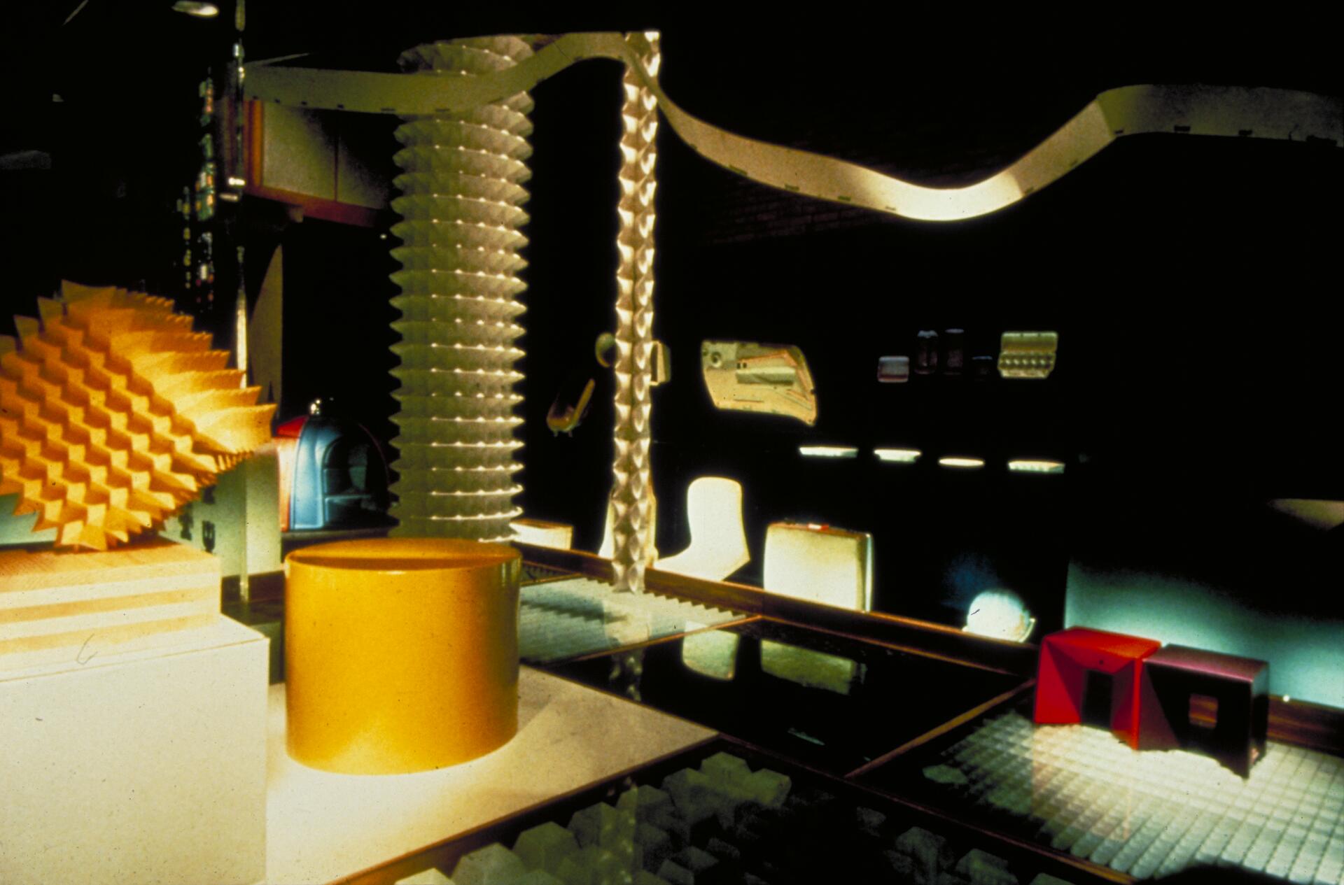 Colorful abstract art installation with geometric shapes in a dimly lit room. Features a large yellow cylinder, a spiraled column, red cubes, and various textured surfaces.