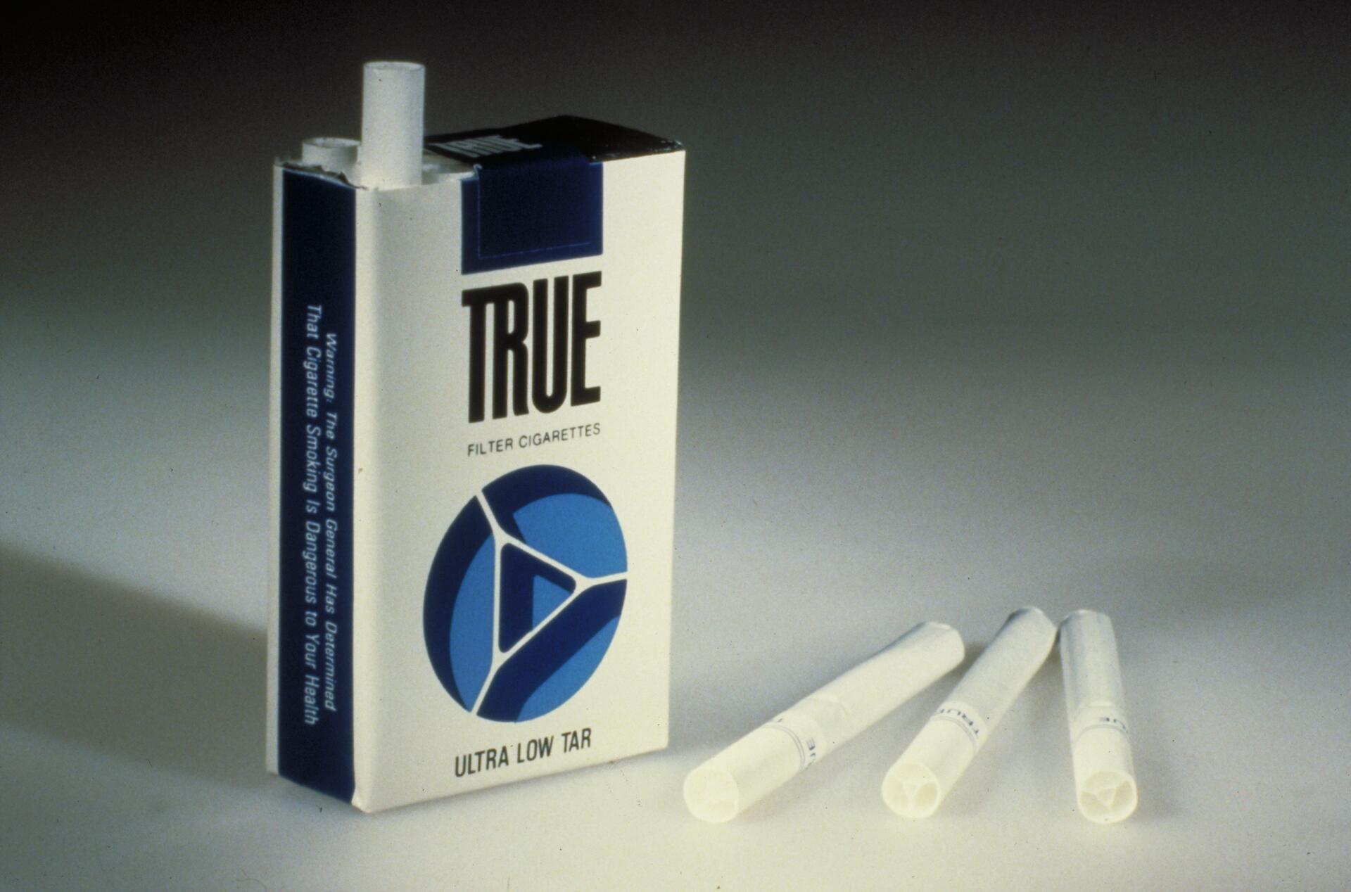 Cigarette pack labeled "TRUE Filter Cigarettes Ultra Low Tar" with two cigarettes lying beside it on a light surface.