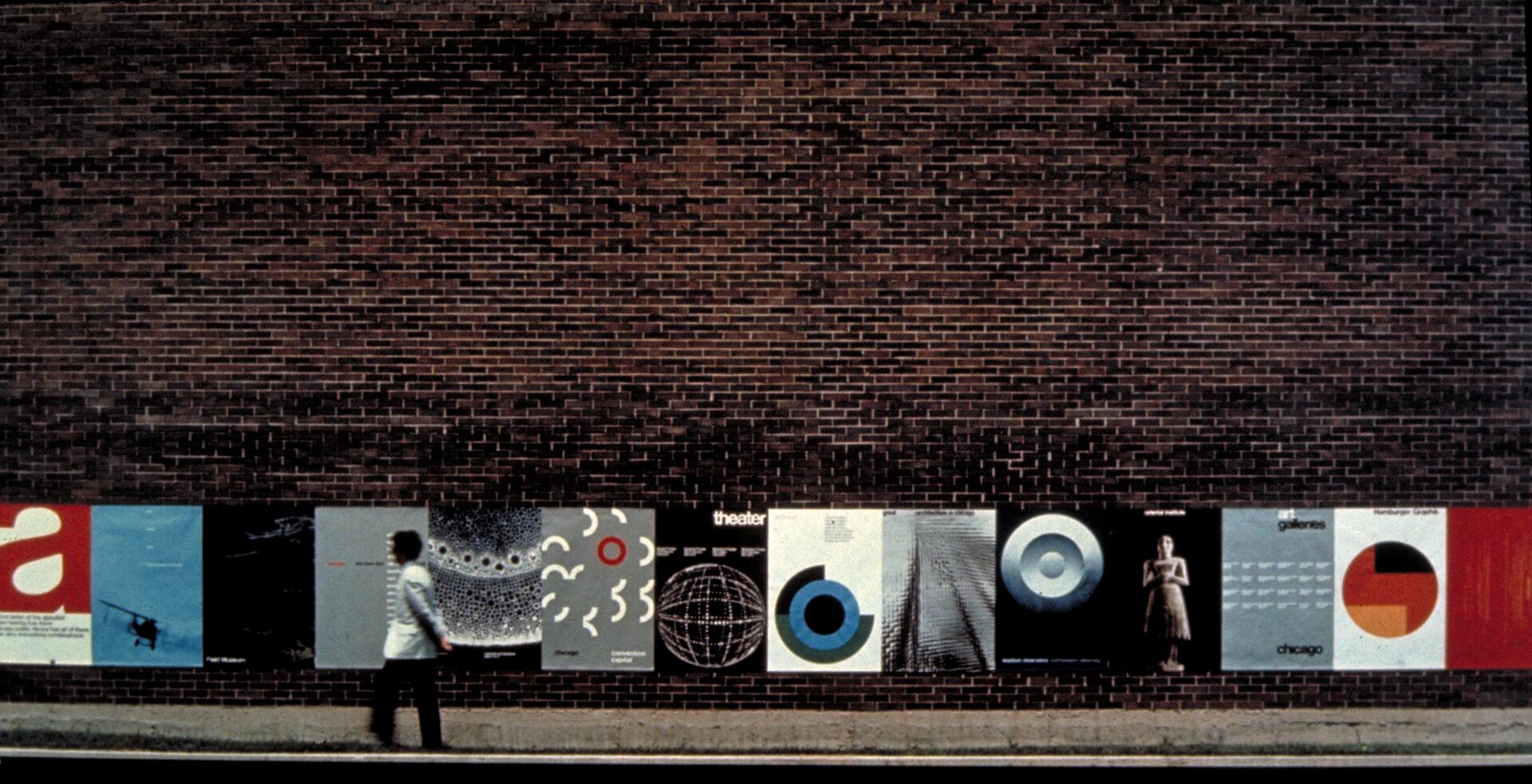 A person walks past posters displayed on a brick wall, showcasing various graphic designs, abstract patterns, and colors.
