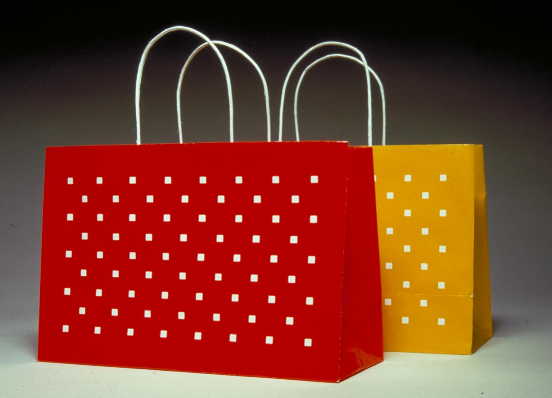 Two paper shopping bags with white square patterns, one red and one yellow, stand side by side against a gradient dark background. Each bag has white handles.