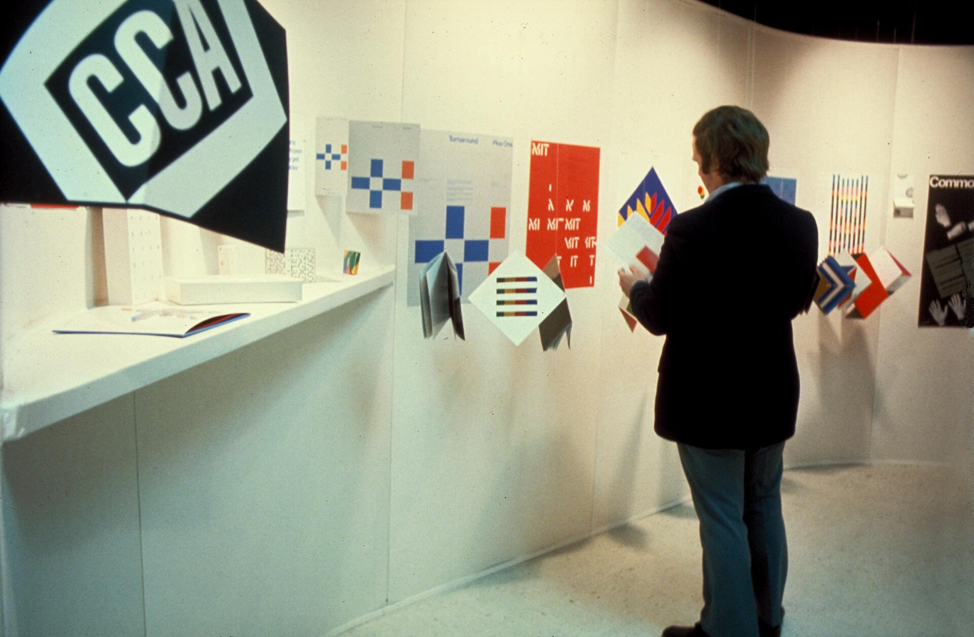 Man viewing a display of colorful graphic design posters on a curved white wall, featuring geometric patterns and bold text.