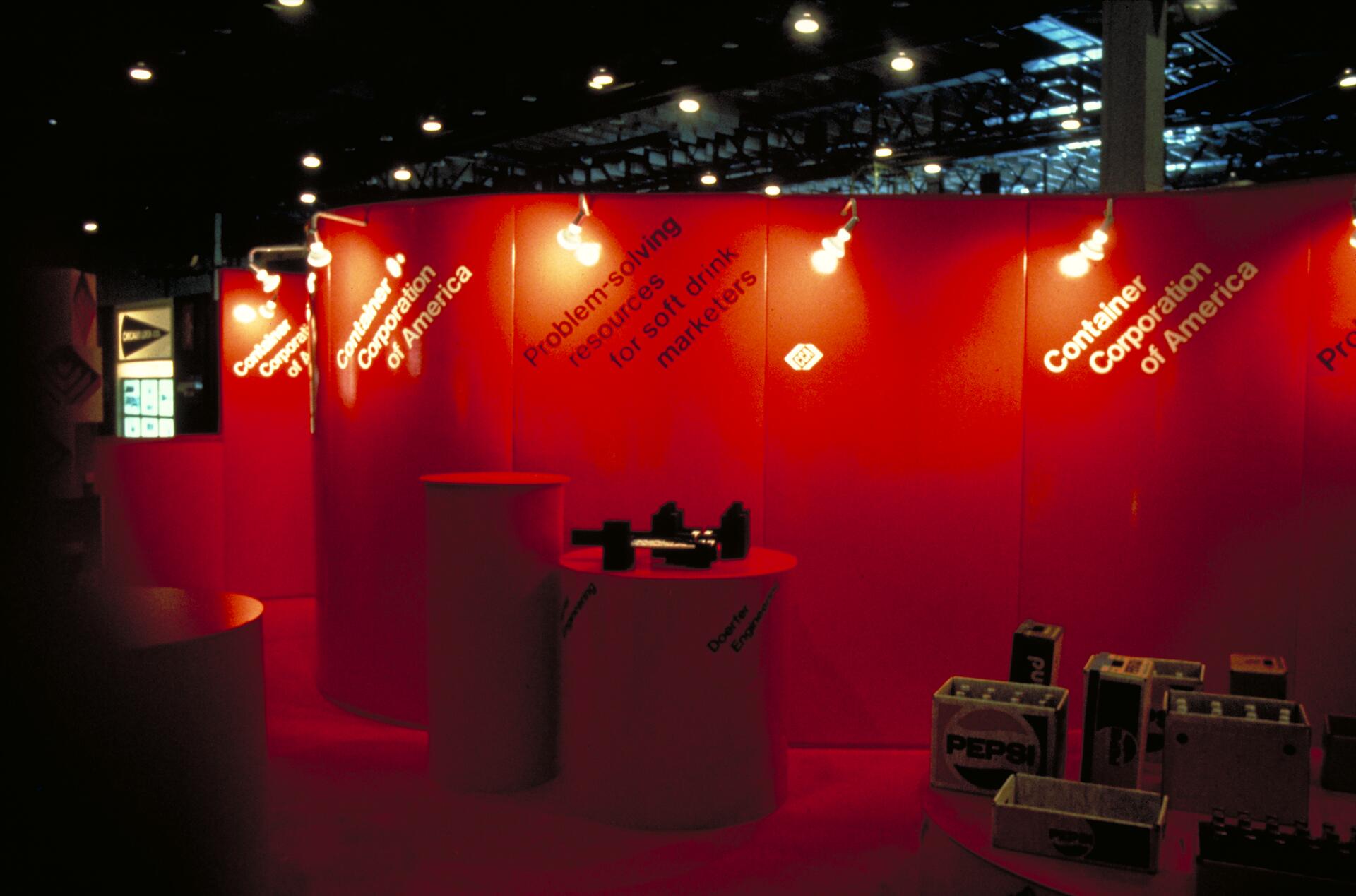 Exhibition booth with red panels and cylindrical display tables. Large white text highlights the Container Corporation of America. Bright lighting and product boxes, including Pepsi-branded items, are visible beneath the spotlights.